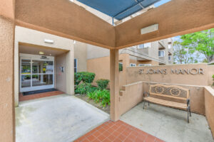 Exterior St. Johns Manor Entrance Signage, bench in front of sign, automatic door at entrance, covered entrance.