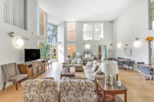 Interior Clubhouse/ leasing office lobby, 2 large couches, contemporary design, white walls, wood floors, 2 lounge chairs, multiple floral paintings, large windows along far-side wall.