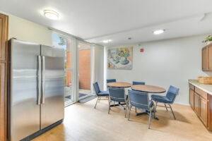 Interior Community kitchen, 2 tables 6 seats, wood floors, brown cabinetry, stainless steel appliances, laminate countertop, glass door to exterior courtyard