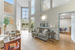 Interior Clubhouse, large contemporary furnishings, multiple 2 person couches, art on the walls, potted plants arranged around the room, large window on the second story with view of the fitness center, adjacent community kitchen to clubhouse, large exit hallway to the courtyard.