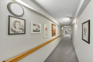 Interior hallway, white walls gray carpeting, portrait and floral art on the walls throughout the hallway.