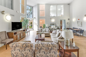 Interior Clubhouse/ leasing office lobby, 2 large couches, contemporary design, white walls, wood floors, 2 lounge chairs, multiple floral paintings, large windows along far-side wall.