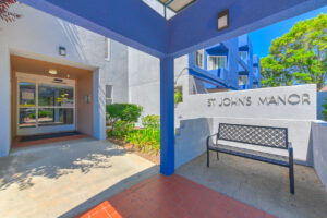 Exterior St. Johns Manor Entrance Signage, bench in front of sign, automatic door at entrance, covered entrance.