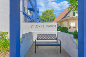 Exterior St. Johns Manor Entrance Signage, bench in front of sign, automatic door at entrance, covered entrance.